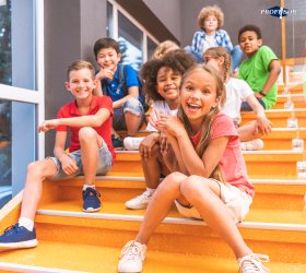 Na zdjęciu widzimy grupę uśmiechniętych dzieci siedzących na pomarańczowych schodach w jasnym wnętrzu. Dzieci wyglądają na szczęśliwe i zrelaksowane, co wskazuje na przyjazną atmosferę panującą w szkole.